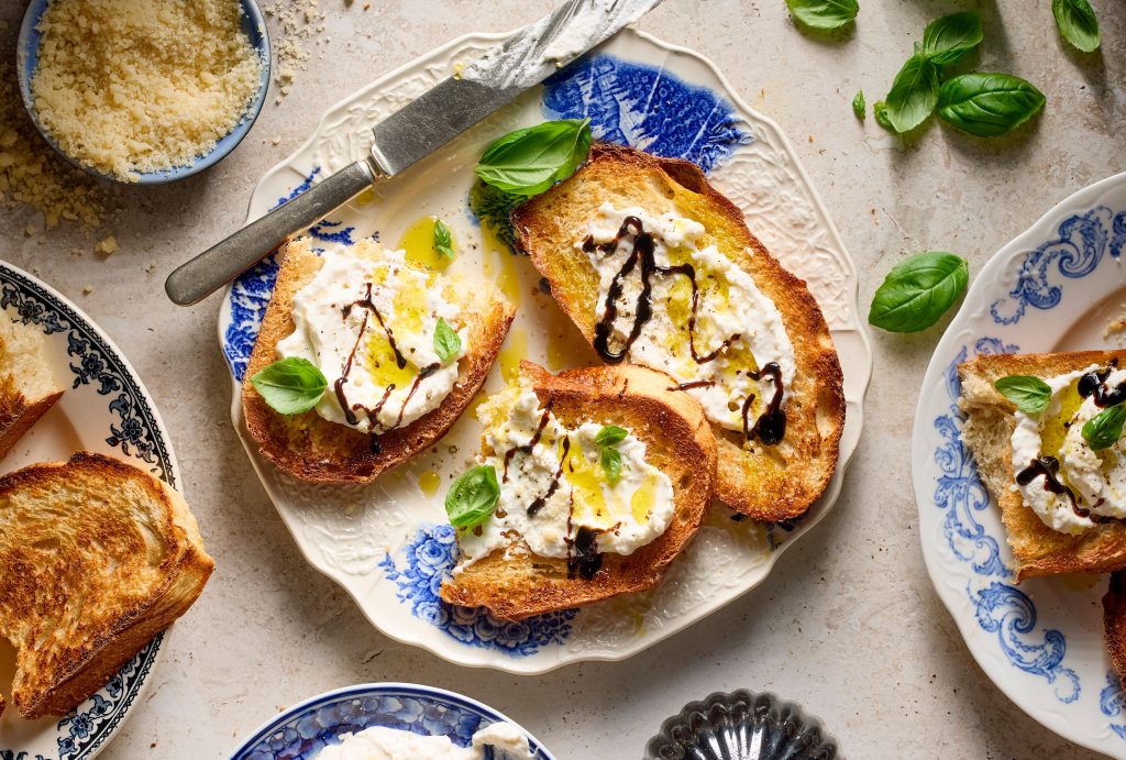 Toasted bread slices topped with a mousse garnished with herbs and a drizzle arranged on a plate with additional garnish nearby