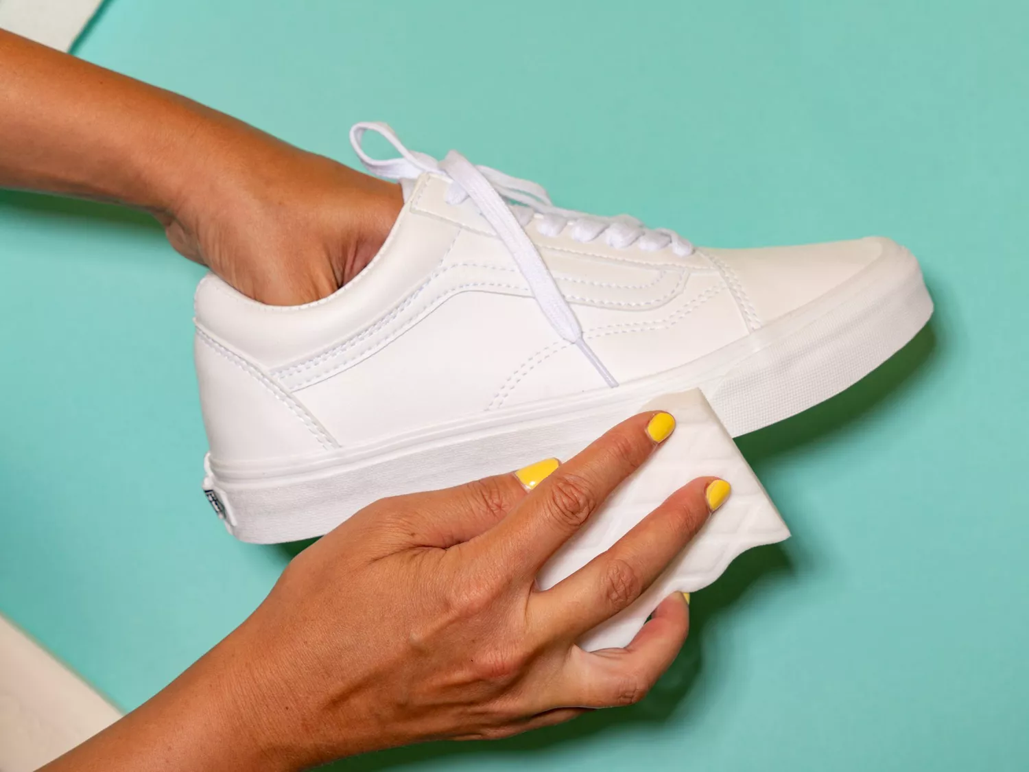 Person using a magic eraser to clean a white canvas shoe, against a teal background