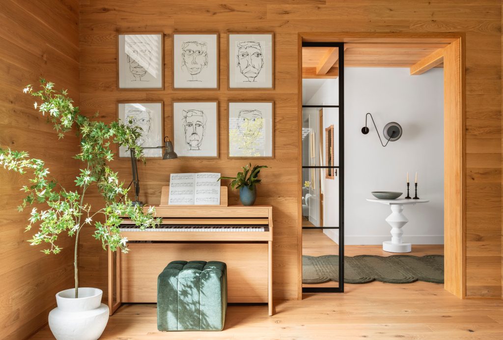 Woodpaneled room featuring a piano with sheet music and decor elements like a plant and wall art leading to a modern styled adjoining space
