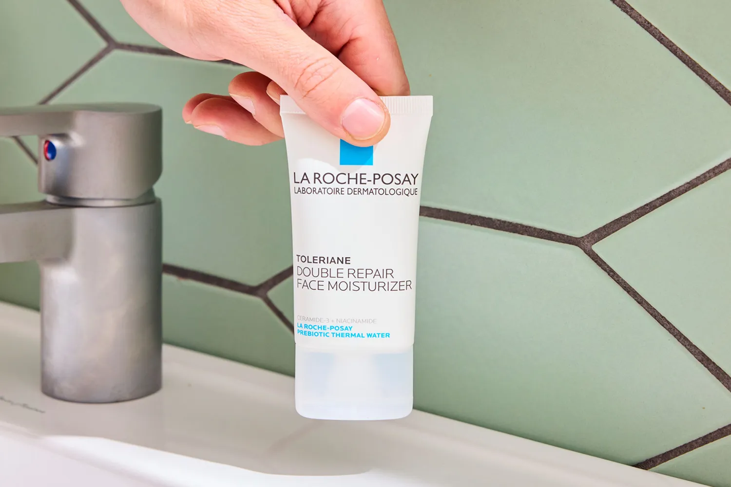 A hand holding a tube of La RochePosay Toleriane Double Repair Face Moisturizer near a sink